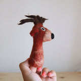 An ethically handcrafted red reindeer wool felt finger puppet is held by a female hand against a plain backdrop, exuding a playful and inviting mood. This charming puppet features a cute red nose, small ears, and brown antlers, all made with a textured wool felt finish, highlighting its unique craftsmanship.