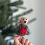 A handcrafted wool felt sea otter with sea star finger puppet is held by a hand against a blurred natural background. This playful sea otter finger puppet features a light tan head, a dark brown body, small ears, a black nose, and black eyes. It holds a vibrant red sea star.