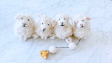 A charming wool felt sheep garland rests on a bright white marble surface. It features three fluffy white sheep, each crafted from wool felt with curly textures, small pink ears, and black stitched eyes and mouths. Black thread connects the sheep to white felt balls, leading to a copper bell at the garland's end.