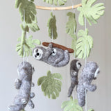 A charming wool felt Sloth Baby Mobile is shown in a close-up against a light wall. It features a wooden frame from which several gray wool felt sloths are suspended by white threads. Each three-dimensional sloth has white facial markings and stitched claws, with wired limbs that can be posed to appear climbing or dangling, evoking a whimsical, handmade charm.