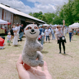 A hand holds a small felt sloth finger puppet, made from wool felt, featuring a brown body with lighter brown spots, a long tail, and a big smile. The background is a rustic setting with a wooden barn and people standing around, creating a casual and relaxed atmosphere, possibly at an outdoor market or fair.