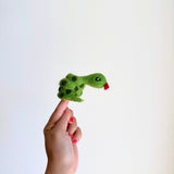 A handmade wool felt snake finger puppet is displayed on a female index finger against a crisp white background. This vibrant green snake puppet features dark green spots, a small red tongue, and tiny eyes. Its coiled shape emphasizes its unique handcrafted design.