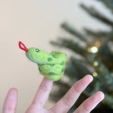 A handmade green snake finger puppet is displayed on a female hand against a soft, neutral backdrop and blurred Christmas tree. This coiled felt finger puppet features green spots, a red tongue, and small eyes, embodying a playful and imaginative spirit for kids' playtime.