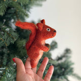 A needle felted squirrel with acorn finger puppet is gently presented by a female hand against a blurred Christmas tree. This charming and realistic squirrel finger puppet is made of textured orange felt, featuring a bushy tail and ears, and holds a small acorn.