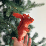 A handcrafted needle felted squirrel with acorn finger puppet is gently held by a female hand against a blurred Christmas tree backdrop. This reddish-brown felted squirrel features fluffy ears and a bushy tail, holding a light brown acorn. Its playful expression adds to its charm.