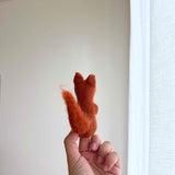 A handmade needle felted squirrel finger puppet is held upright in a female hand against a soft white background near a window with sheer white curtains. This reddish-brown wool felt squirrel puppet features a textured appearance, a bushy tail, small ears, and a rounded body, showcasing its detailed design and craftsmanship.