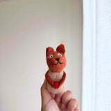 A handmade needle felted squirrel finger puppet, ethically crafted by female artisans, is held up by a female hand against a bright, neutral background. This charming squirrel puppet features a reddish-brown head and arms, a cream-colored body and ears, a stitched smile, and holds a small red acorn.