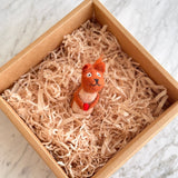 A handcrafted needle felted squirrel with acorn finger puppet rests inside a square cardboard box filled with light shredded paper, set on a white marble surface. The orange and cream squirrel features a black nose and mouth, holding a small red acorn, and conveys a curious, playful expression. This unique felt toy for kids offers a cozy, natural feel.
