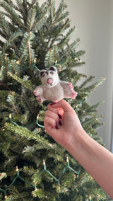 A festive needle felted sugar glider finger puppet is held by a hand, set against a twinkling Christmas tree. This handcrafted wool felt sugar glider is a 3D design in light gray, with darker gray ears, a pink nose, and pink paws. Its unique and cheerful presence enhances the cozy, playful holiday atmosphere.
