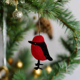 A red Taiwanese Po-Yo bird felt ornament hangs from a deep green Christmas tree branch. The plump bird features a black beak, black feet, and a black marking across its face and down its chest, suspended by a gold string. Blurred gold ornaments add a warm glow in the background.