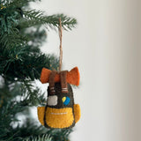 This handcrafted yellow and black wool felt travel backpack ornament is displayed on a green Christmas tree. It features an orange rolled sleeping pad strapped to the top with brown felt straps, and a grey square with a blue and yellow circle design. A jute string is attached for hanging.