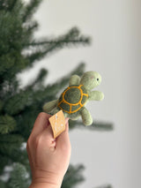 A handcrafted wool felt turtle finger puppet is presented, held by a female hand, with a blurred Christmas tree in the background. This unique felt turtle finger puppet is light green with a darker green shell outlined in yellow. It features small, rounded flippers and a sweet expression, reflecting its playful and artisan-made design.
