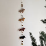 A felt Dachshund dog garland hangs on a plain white wall, adding a playful and festive touch. It features four evenly spaced felt Dachshunds, each in a different color: cream, khaki, brown, and black. A blurred evergreen branch is visible on the right side.