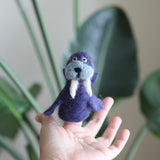 A handmade wool felt walrus finger puppet is displayed on a fingertip by a female hand, against a blurred backdrop of green foliage. This unique, needle-felted walrus puppet is purple, featuring flippers, light blue around its mouth, and distinct white tusks. Crafted from 100% wool, it represents a charming and environmentally friendly toy.