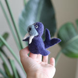 A handcrafted needle-felted wool walrus finger puppet is gently held by a hand against a soft, blurred background of large green leaves. This unique purple walrus puppet, ethically handmade in Nepal, features a light blue face, prominent white tusks, black eyes, and a small tail, perfect for imaginative play.