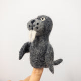 A handmade gray wool felt walrus finger puppet is shown on a finger against a plain white background, demonstrating its playful use. This unique needle-felted walrus puppet, crafted from 100% wool, features a rounded, 3D body with small flippers, a tail, and a slightly fuzzy texture. Its charming face includes dark button-like features for the nose and mouth, complemented by white felt tusks.