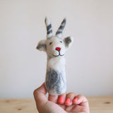 A handmade needle felted white goat finger puppet is held by a female hand with pink nail polish, presented against a soft, neutral backdrop. Crafted from wool felt, this unique goat puppet features a white body with gray and black markings, gray ears, and gray and white striped horns. Its friendly face has a red nose and a stitched black smile, highlighting its fine, handcrafted details.