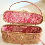 A small, round basket made from woven rattan with a floral pattern sits on a wooden table. The rattan bag features a handle at the top for carrying and is open, revealing its interior lined with Batik cotton fabric.