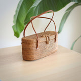 A woven basket with a handle, resting on a table surface made from natural rattan material, featuring a simple design without patterns or embellishments. The brown leather handle contrasts with the lighter color of the woven material. A plant with broad leaves is visible in the background, adding greenery to the scene.