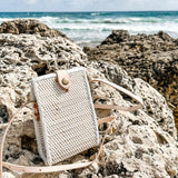 A beige woven rattan crossbody cellphone bag rests on a rocky surface near the ocean, surrounded by clear blue skies and calm waters. The bag features a high-quality woven rattan material with a natural texture, an adjustable strap made from real leather, and a secure snap closure. The lining is made of unique Batik cotton, adding to both style and durability.