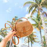 A handheld crossbody bag made from natural materials resembling Ata grass, adorned with a batik pattern resembling palm leaves. The bag features a real rattan strap that is adjustable and has a snap closure, resting on the arm of a person in the background who is holding it.
