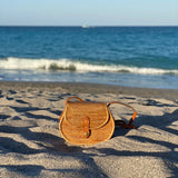 A Bali Rattan Crossbody Bag rests on a sandy beach near the ocean, showcasing its natural earthy color tone and woven rattan material. The bag features a rectangular shape with a snap closure and adjustable strap for a customized fit.