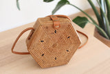 A woven rattan crossbody bag, Bali Hexagon Rattan Crossbody Bag - Hexagon, rests on a wooden surface. The hexagonal-shaped bag features a geometric design created by intersecting lines and an adjustable brown leather strap. A plant and white wall are visible in the background, suggesting an indoor environment.