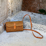 A small crossbody bag made from natural materials, likely rattan or bamboo, rests on a stone surface against a tropical backdrop featuring a wall and foliage. The rectangular-shaped bag with rounded corners showcases a woven texture in a light brown color, adorned with a small round leather detail on one side. Its adjustable strap hangs slightly downwards, blending seamlessly into the casual outdoor setting.
