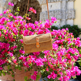 A small, woven rattan crossbody bag with a leather strap hangs from it, displayed in front of a vibrant flowering bush. The handcrafted bag features intricate weaving patterns and is set against the backdrop of a stone building with an archway, evoking a relaxed and leisurely mood. The product's design and material suggest casual outings or fashionable accessories for warm weather climates.