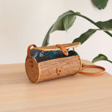 A woven crossbody bag, Bali Rattan Crossbody Bag - Tapang, rests on a surface. The cylindrical-shaped bag features a textured weave pattern with a bohemian look, showcasing its eco-friendly and fair-trade design. A real leather strap is adjustable for comfort, and the bag's interior boasts a beautiful batik lining.