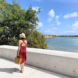 A woman walks along a paved pathway next to a body of water, carrying a large rattan handbag featuring a butterfly pattern. The handbag is made from wicker or rattan material with real rattan handles and a unique butterfly wicker design.