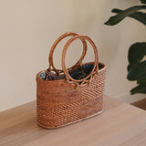 A woven rattan handbag, Bali Rattan Handbag - Kelsey, rests on a surface. The bag is made from natural materials like ata grass for the circular handles, giving it a textured appearance. This stylish yet functional accessory features a spacious interior and a batik lining drawstring pouch.