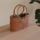 A woven basket with a handle on top, resting on a wooden surface. The basket is crafted from rattan or similar natural material, featuring a textured appearance and colorful fabric or thread accents. Its eco-friendly design showcases the beauty of handmade craftsmanship.