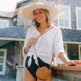 A woman stands outdoors, wearing a white top, black shorts, and a straw hat. In front of her, a wooden table holds a small purse. The background features a house with a shingled roof and deck area. A Bali Round Mini Rattan Crossbody Bag sits on the table, showcasing its natural rattan material and unique Batik lining.