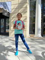 A young girl stands on a sidewalk next to a building, holding a book titled 'Mia' and wearing a blue shirt with a graphic print. She holds the Bali Round Mini Rattan Crossbody Bag - Mia, crafted from natural rattan with a vintage aesthetic.