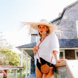 A woman stands outdoors, wearing a straw hat and holding our Bali Round Mini Rattan Crossbody Bag. The bag is crafted from natural rattan with a vintage aesthetic, featuring a round shape, earthy tones, and a woven texture. A unique Batik lining adds an extra touch of style.