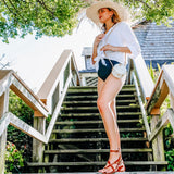 A woman stands on a wooden staircase, wearing a white top and straw hat, holding a small round rattan bag. The bag's natural texture and boho-chic detailing are showcased, with its shape and material aligning with its description as a crossbody or shoulder bag.