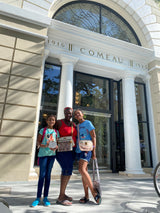 A casual outdoor scene featuring three individuals posing on a sidewalk in front of an arched doorway building, with one person holding the Bali Round Mini Rattan Crossbody Bag - Mia. The bag is crafted from natural rattan with a vintage aesthetic, designed for convenience and practicality.