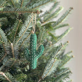A wool felt cactus ornament hangs on a Christmas tree, creating a festive and natural atmosphere. The green cactus features white stitching, adding a textured detail to its design. A hemp string is attached for easy hanging, visible against the green needles of the tree.