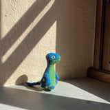 A handcrafted wool felt Brontosaurus finger puppet stands on a light gray surface near a dark wooden frame. This playful needle-felted Dino Brothers felt toy features a long neck and majestic shape, adorned with a vibrant mix of blue and green and white spots. The charming scene is set against a neutral wall, illuminated by soft sunlight casting gentle shadows.