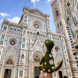 A green wool felt Brontosaurus finger puppet, a unique felt toy for kids, is held on a person's finger against a bright sky and the ornate facade of a cathedral. This handcrafted, needle-felted dinosaur features a long neck, a long tail, and small white spots. Handmade in Nepal from 100% wool, it offers a lifelike appearance for imaginative play.
