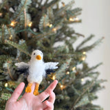 A handcrafted wool felt eagle finger puppet is gently held by a female hand, presented against a softly lit Christmas tree backdrop. This unique needle felted eagle, with its iconic fluffy hair and charming aesthetic, is ready to inspire festive storytelling and imaginative play.