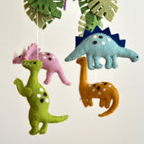 A vibrant felt baby mobile, featuring Jurassic dinosaurs, hangs against a calming off-white background. The white felt frame suspends colorful felt dinosaurs, including a green T-Rex, pink Triceratops, blue Stegosaurus with dark blue plates, orange Brontosaurus, and a Pterosaur, alongside green monstera leaves. Each dinosaur is crafted with charming details and stitched accents, creating a playful and soothing display.