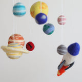 A colorful wool felt baby mobile, featuring planets and a spaceship, hangs from a string against a white background. The mobile is handcrafted with intricate details and textures, creating a sense of balance and harmony. Each planet has its own distinct color and design, representing various celestial bodies in our solar system. This unique baby mobile is made from soft, colorful wool felt, perfect for inspiring curiosity about space and the universe in children's rooms.