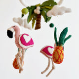A colorful felt baby mobile, titled 'Tropical Trio', featuring three vibrant elements: a pink flamingo on the left, a pineapple and palm tree on the right. The mobile is suspended from a string attached to a hook, with a neutral background allowing the bold colors of the felt figures to stand out.