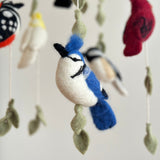 A serene and playful felt baby mobile, Wings of America, hangs from a string, featuring various birds like blue jays, hummingbirds, and chickadees decorated with leaves on a wooden frame in an indoor setting.