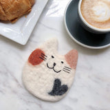 A round felt calico cat coaster, part of a set, rests on a white marble surface beside a coffee cup and a croissant, creating a cozy, inviting scene. This handcrafted wool felt coaster, ethically made by Nepalese female artisans, features a distinct calico pattern with white, orange, and pink ears, a black spot, and needle felted whiskers and smile. It provides a whimsical touch and protects surfaces.