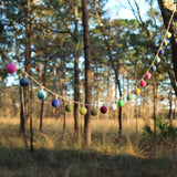 A colorful wool felt lightbulb garland is strung between two trees, set against a natural outdoor backdrop of green foliage. The garland features numerous soft, round felt balls, needle-felted from 100% wool, in shades of pink, blue, green, yellow, white, and maroon. Each colorful felt ball hangs individually, creating a whimsical and handmade feel.
