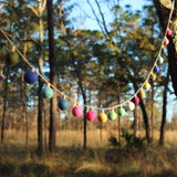 A colorful felt lightbulb garland, featuring individual 3D lightbulb shapes in vibrant blue, pink, green, white, yellow, and brown, is strung between two trees. Made from 100% wool felt, the garland hangs on a thin, light-brown string, set against a blurred outdoor backdrop of natural foliage on a sunny day.