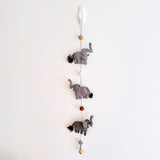 A felt elephant garland is suspended from a white hook against a plain white wall. The garland features three gray felt elephants, each detailed with white tusks and black yarn tails. Round felted balls in brown and white separate the elephants. A small copper bell is attached at the bottom.